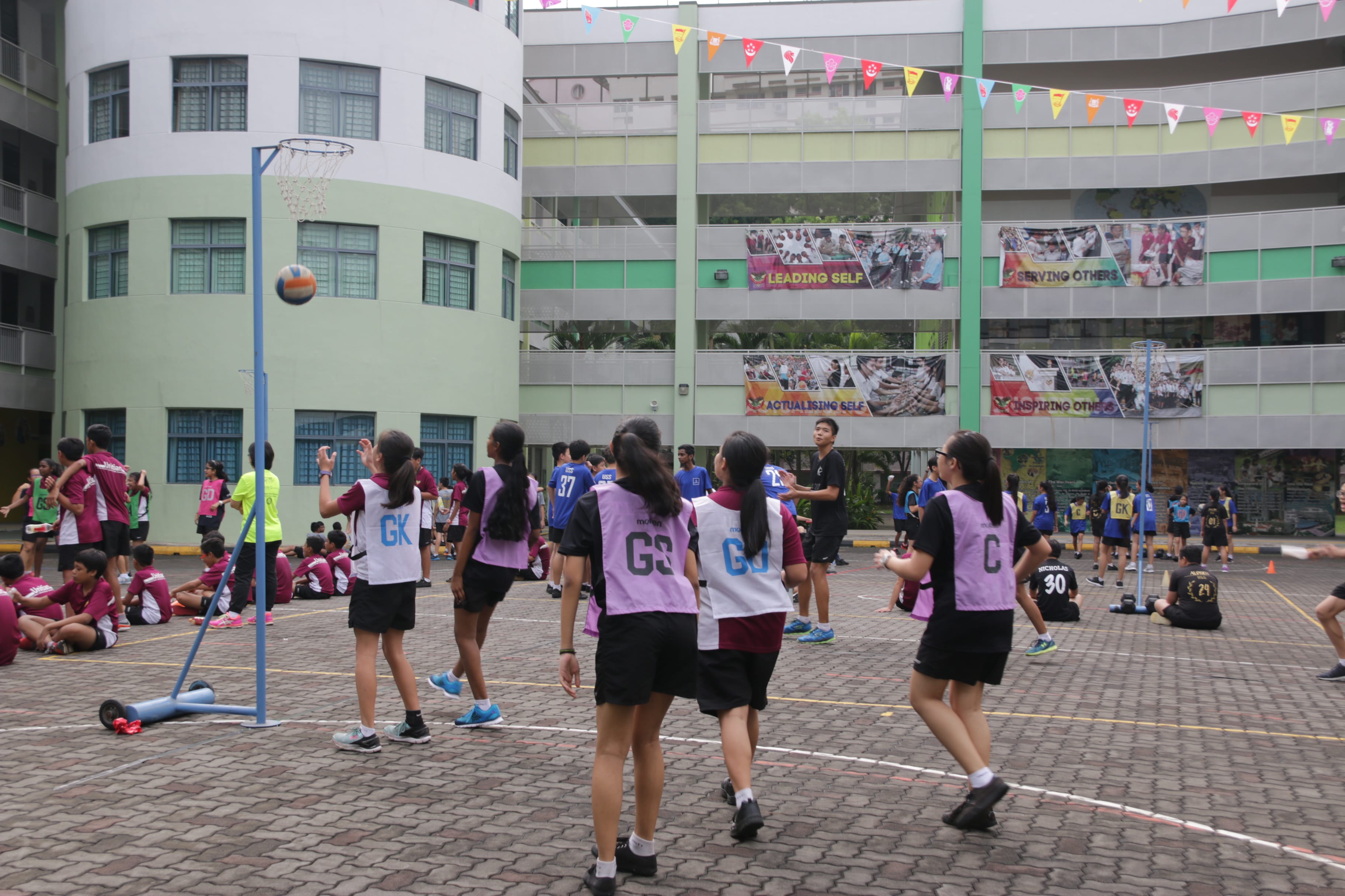 Students in purple bibs play netball on an outdoor school court with a ball in the air near the post.